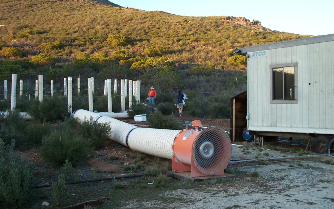 Scientific infrastructure on a hillside: a ring of vertical white tubes and a larger, horzontal white pipe with a fan